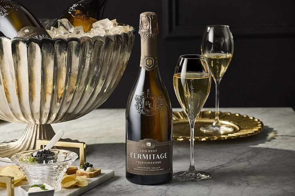 A bottle of L'Ermitage 2019 Brut is on a table with a champagne flute. Nearby are potato chips and a plate of caviar and garnishes on ice.