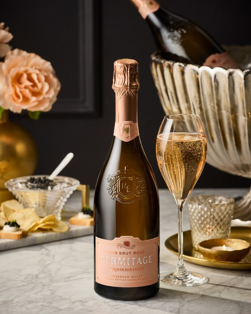 A bottle of 2019 Brut Rosé sparkling wine stands on a marble surface. A filled glass is beside it. In the background, there are peach roses, a silver ice bucket with another bottle, and snacks like chips and caviar. The ambiance is elegant and luxurious.