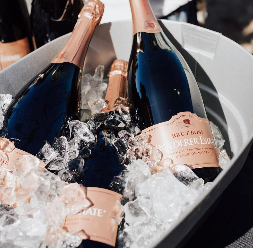 Bottles of Brut Rosé champagne are chilled in a tub filled with ice. The labels read "Roederer Estate" and "Anderson Valley." The bottles have a pink foil on the neck, and sunlight reflects off the ice and glass.