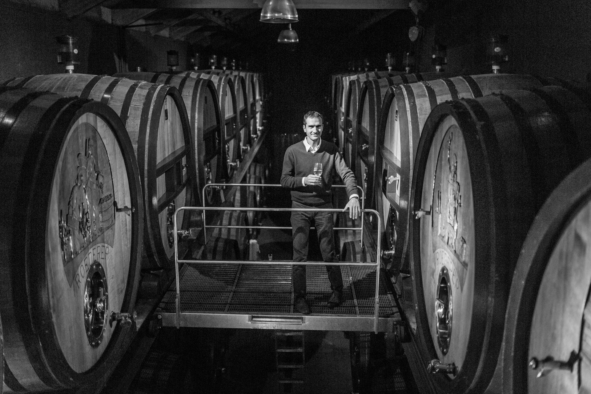 A person stands on a metal walkway in a dimly lit winery, holding a wine glass. Surrounding them are large wooden barrels lined up on either side.