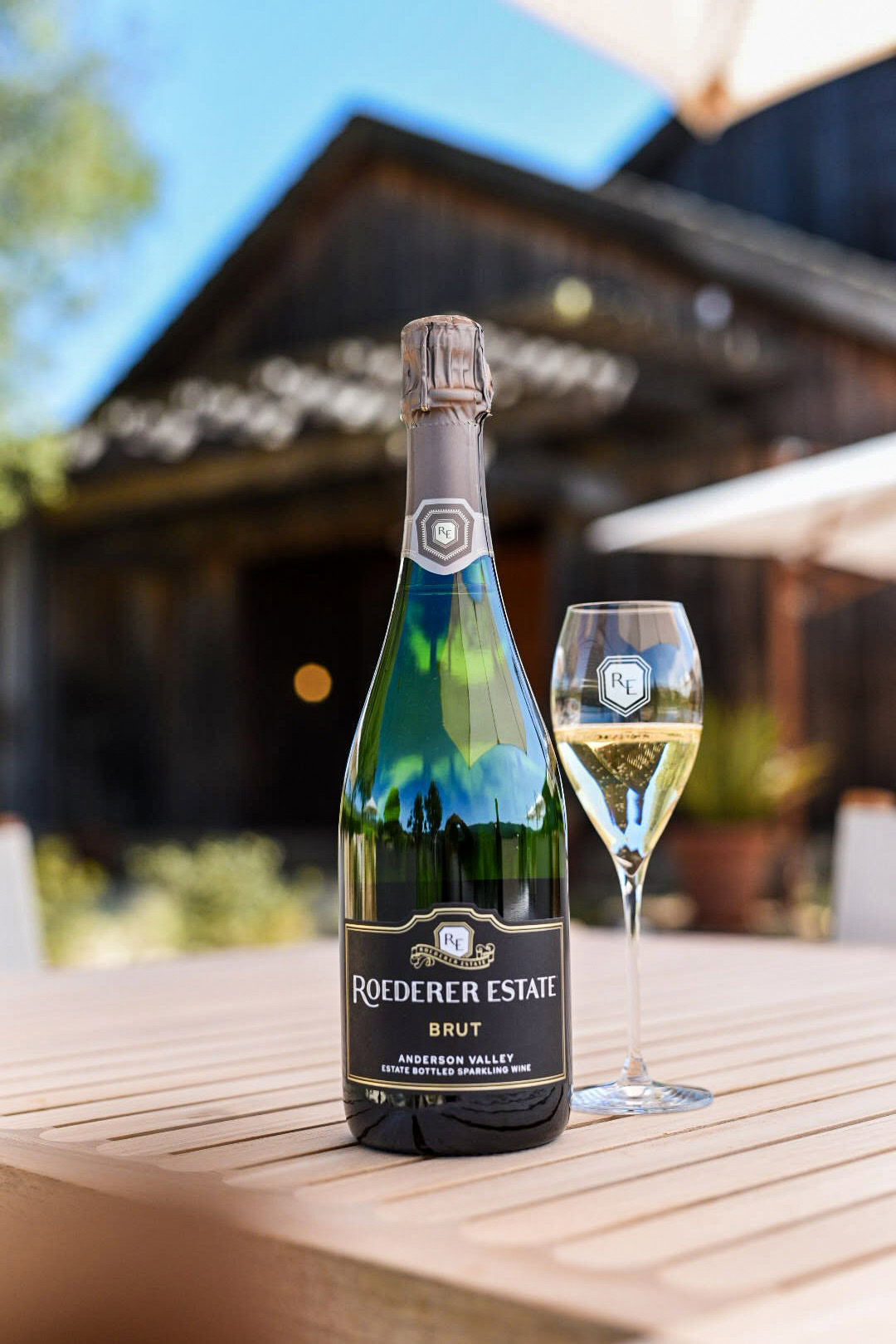 A bottle of Roederer Estate Brut and a filled champagne flute are on a wooden table outdoors. A rustic wooden building and a plant are in the blurred background under a sunny sky.