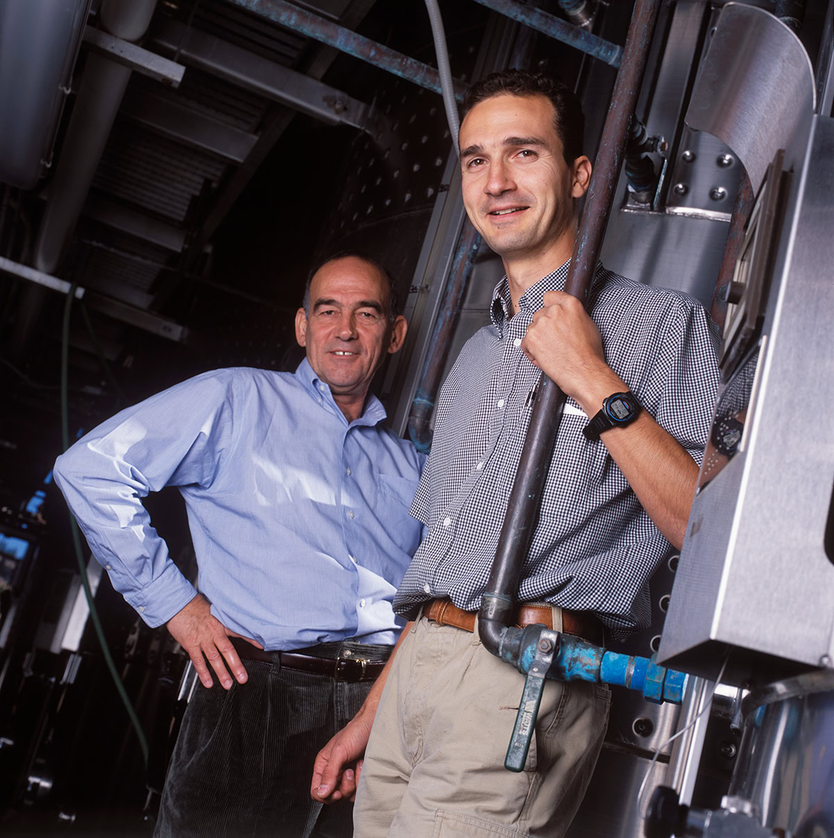 Two men standing in a winery setting, surrounded by machinery. One is wearing a light blue shirt and dark pants, the other a checkered shirt and khaki pants. Both are smiling and looking at the camera.