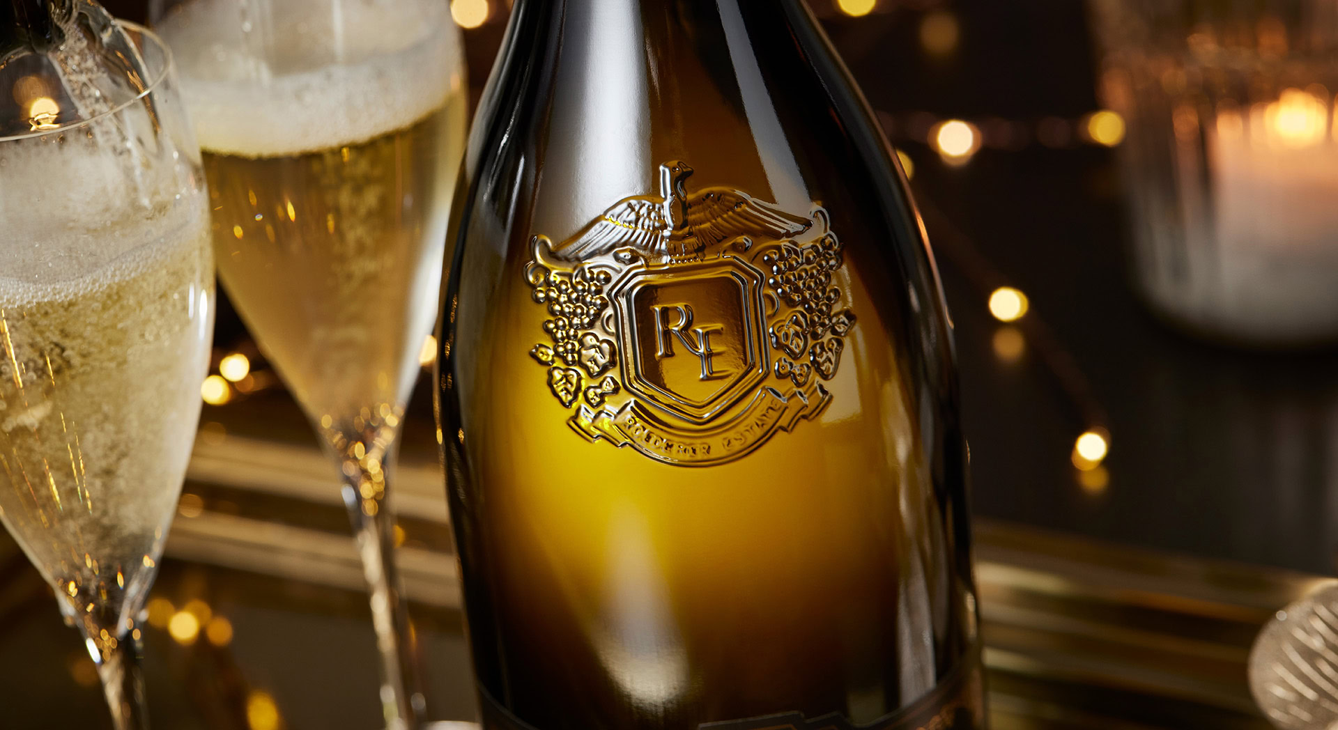 Close-up of an ornate wine bottle with a raised emblem featuring the letters "RP." Two filled champagne glasses are beside it. Soft, warm lighting and blurred golden fairy lights create an elegant ambiance.