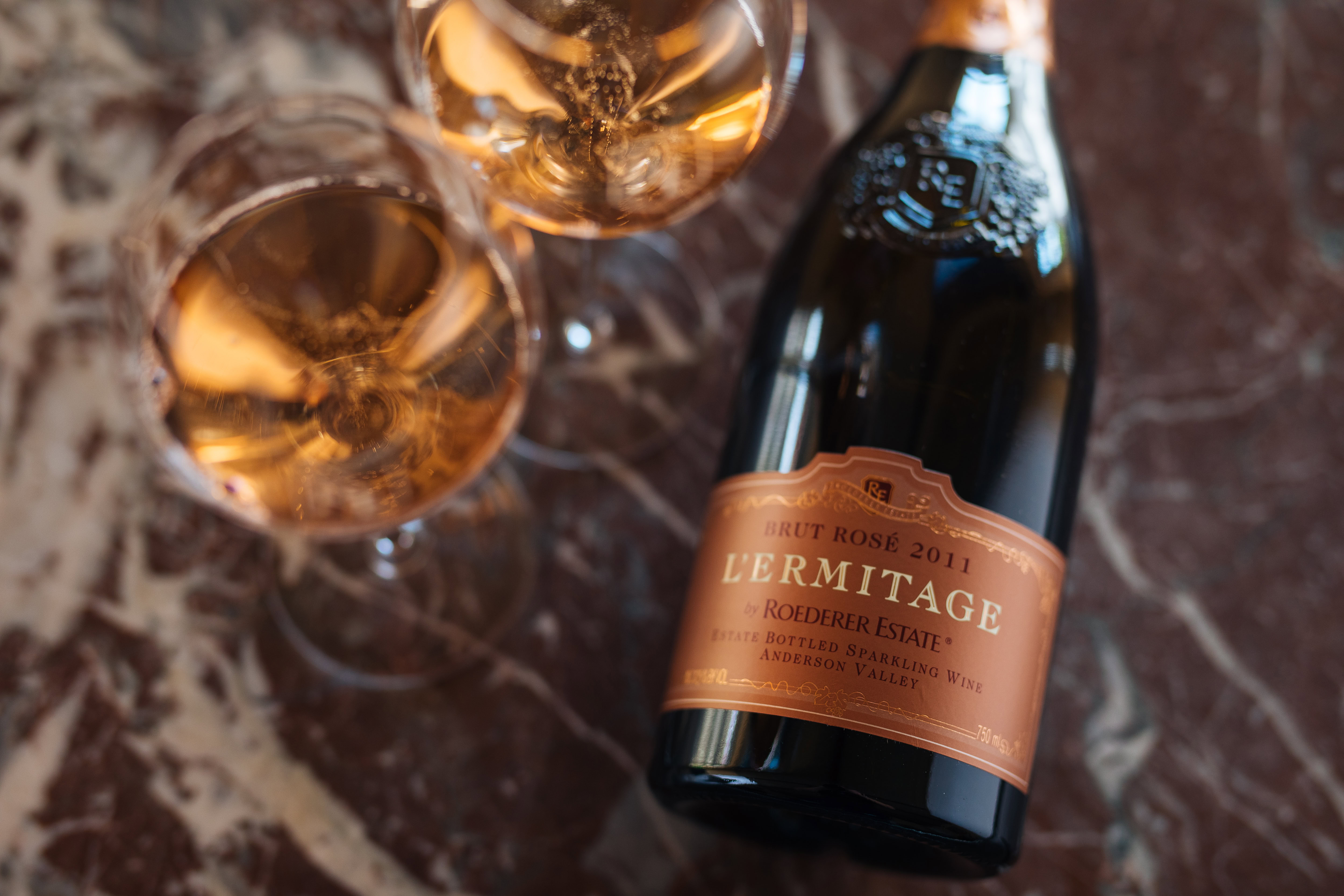 Two glasses of sparkling rosé wine are filled near a bottle of Brut Rosé 2011 L'Ermitage by Roederer Estate. The bottle and glasses are placed on a marble surface.