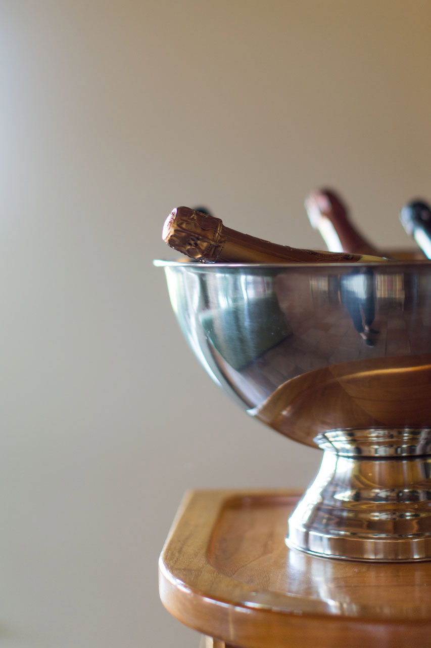 A silver ice bucket holding several bottles of champagne sits on a wooden table. The corks are wrapped in foil and visible. The background is softly blurred, emphasizing the curved reflection of the room on the metal surface.