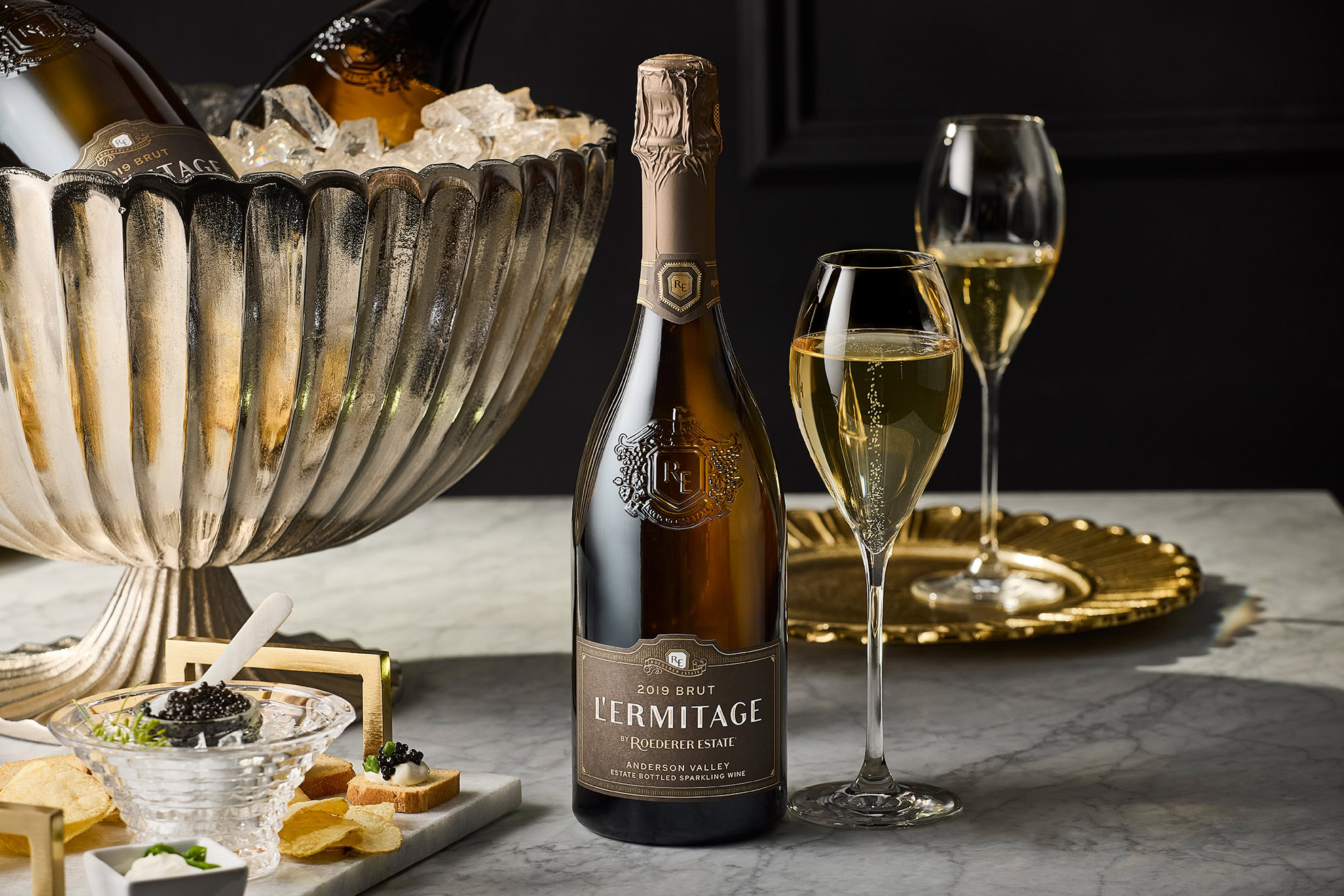 Bottle of L'Ermitage sparkling wine on a marble table with a filled glass next to it. Ice bucket with another wine bottle and a glass being poured in the background. Caviar and breadsticks on a plate. Elegant, dark setting.