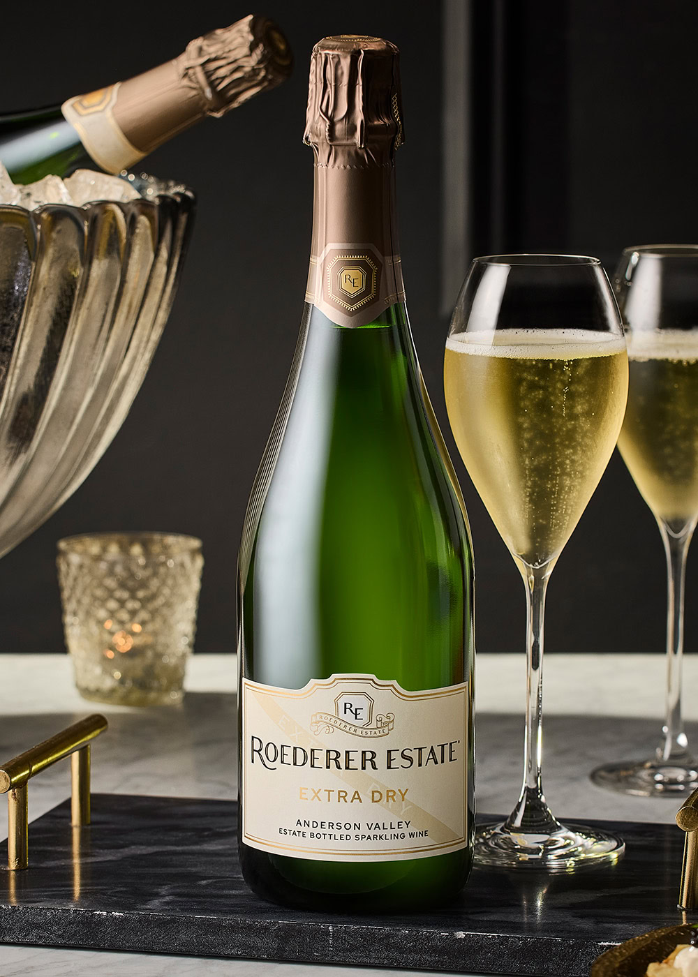 Bottle of Extra Dry sparkling wine on a marble table with a filled glass next to it. Ice bucket with another wine bottle and a glass being poured in the background. Elegant, dark setting.